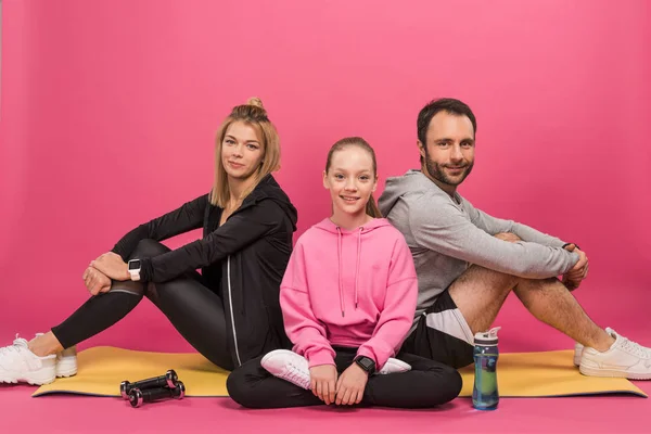 Familia atlética sentada en una colchoneta de fitness con mancuernas y botella deportiva, aislada en rosa — Stock Photo