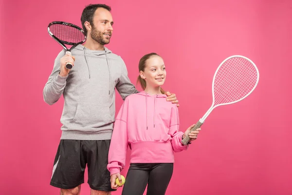 Спортивный отец и дочь-подросток, держащие теннисные ракетки и мяч, изолированные на розовый — стоковое фото