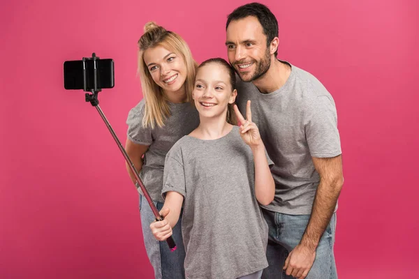 Sonriente familia tomando selfie en smartphone mientras hija mostrando símbolo de paz, aislado en rosa - foto de stock