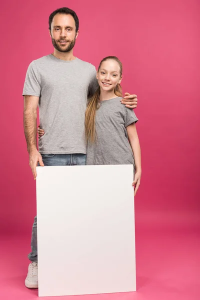 Батько обіймає дочку і позує з порожнім плакатом, ізольовано на рожевому — стокове фото