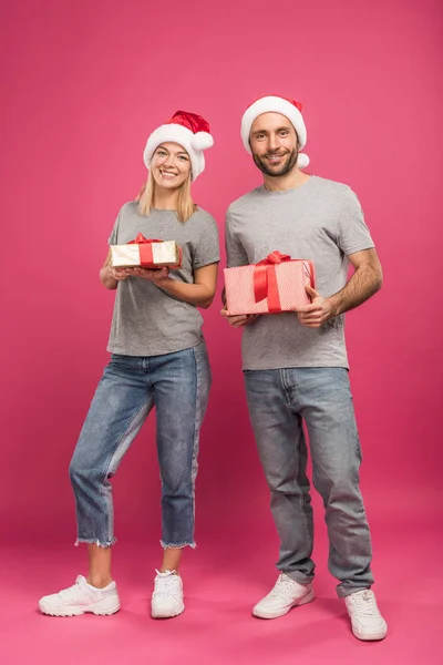 Happy couple in santa hats holding christmas presents on pink — Stock Photo