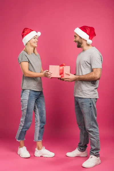 Beautiful happy couple in santa hats gifting christmas present on pink — Stock Photo