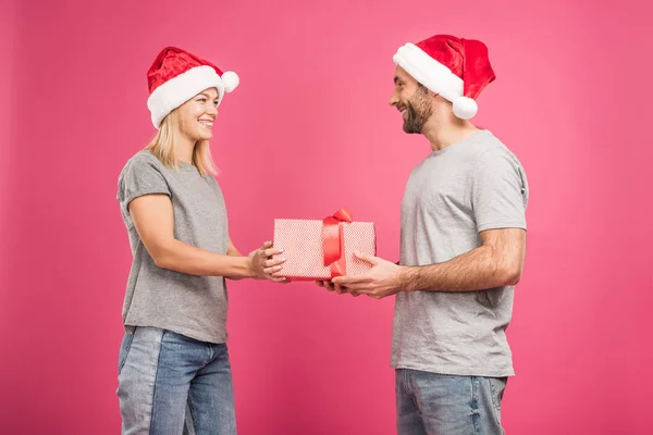 Beautiful cheerful couple in santa hats gifting christmas present, isolated on pink — Stock Photo