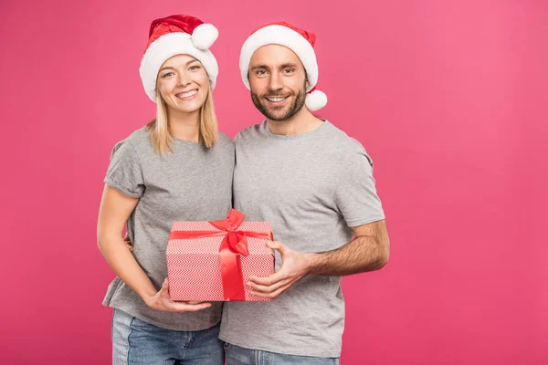 Sorrindo casal em chapéus de santa abraçando e segurando caixa de presente de Natal, isolado em rosa — Fotografia de Stock