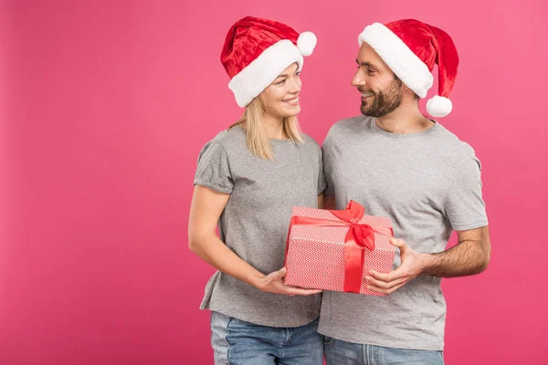 Happy couple in santa hats hugging and holding christmas gift box, isolated on pink — Stock Photo