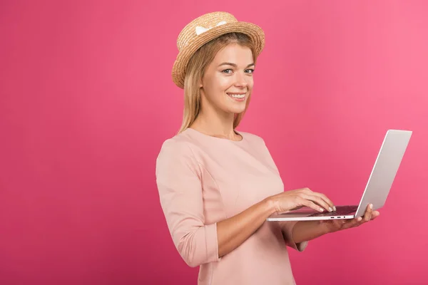 Atractiva mujer alegre en sombrero de paja usando el ordenador portátil, aislado en rosa - foto de stock