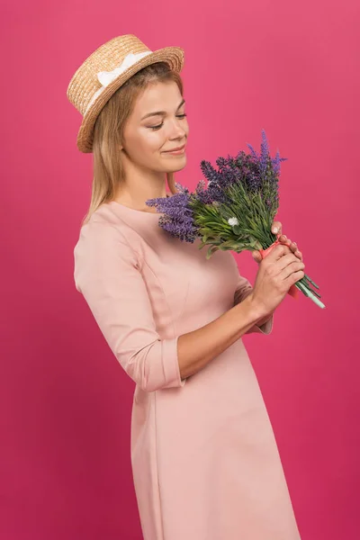 Happy woman in straw hat looking at bouquet of flowers, isolated on pink — Stock Photo