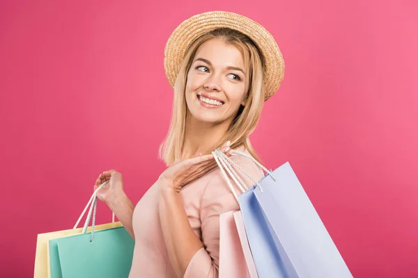 Mulher sorridente atraente em chapéu de palha segurando sacos de compras, isolado em rosa — Fotografia de Stock