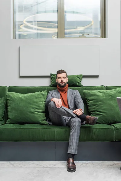 Uomo in abito formale con le gambe incrociate seduto sul divano verde e guardando la fotocamera — Foto stock