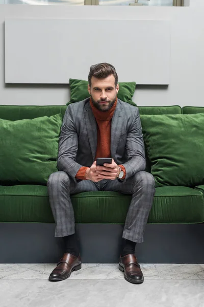 Целенаправленный красивый мужчина в формальной одежде сидит на зеленом диване, смотрит в камеру и использует смартфон — стоковое фото