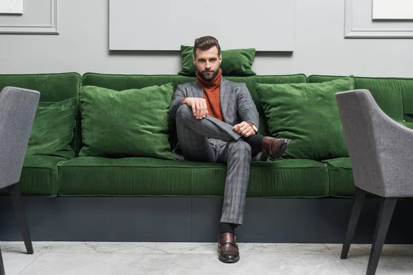 Мужчина в формальной одежде со скрещенными ногами сидит на зеленом диване и смотрит в камеру — стоковое фото