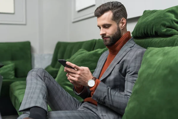 Focused handsome man in formal wear sitting on green sofa and using smartphone — Stock Photo