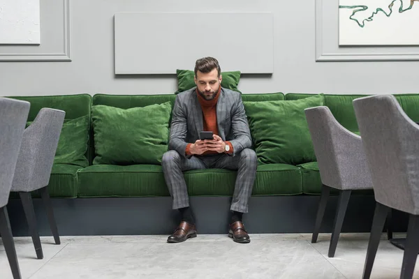 Hombre guapo serio en ropa formal sentado en el sofá verde y el uso de teléfono inteligente - foto de stock