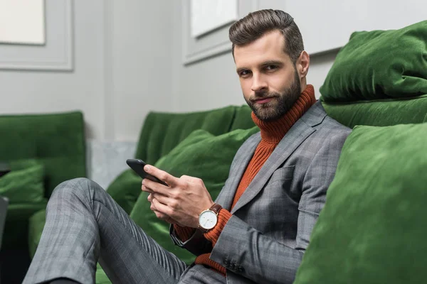 Homem bonito em desgaste formal sentado no sofá verde, olhando para a câmera e usando smartphone — Fotografia de Stock