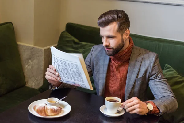 Guapo hombre elegante en ropa formal sentado en la mesa durante el almuerzo y la lectura de periódicos en el restaurante - foto de stock