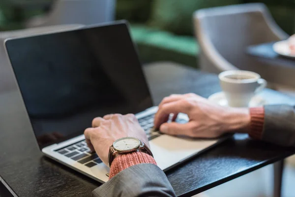 Cropped view of businessman sitting at table and typing on laptop with blank screen in restaurant — Stock Photo