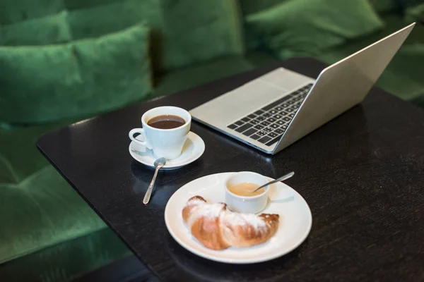 Foco selectivo de plato con croissant y salsa, café y portátil en la mesa en la cafetería - foto de stock