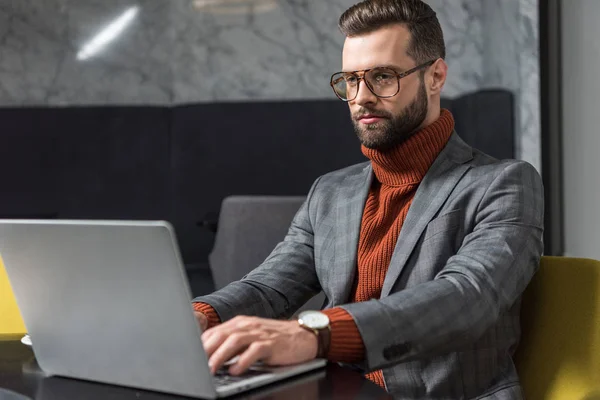 Handsome businessman in formal wear and glasses sitting at table and typing on laptop in restaurant — Stock Photo