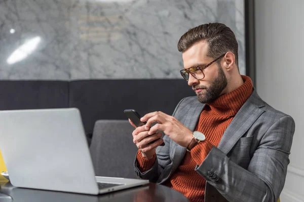 Handsome businessman using smartphone and sitting at table with laptop in restaurant — Stock Photo