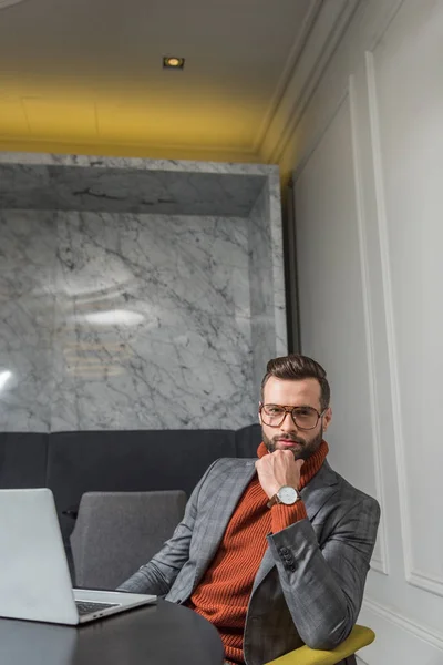 Guapo hombre de negocios en ropa formal sentado en la mesa con el ordenador portátil en el restaurante y mirando a la cámara - foto de stock