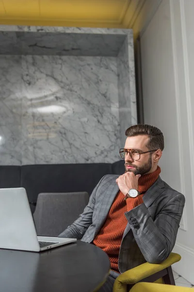 Задумчивый привлекательный бизнесмен в формальной одежде сидит за столом с ноутбуком в ресторане — стоковое фото