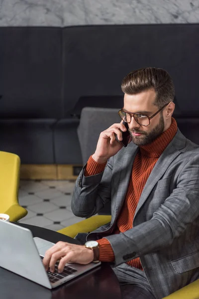 Серьезный красивый бизнесмен в очках и формальной одежде сидит за столом, разговаривает на смартфоне и печатает на ноутбуке в ресторане — стоковое фото