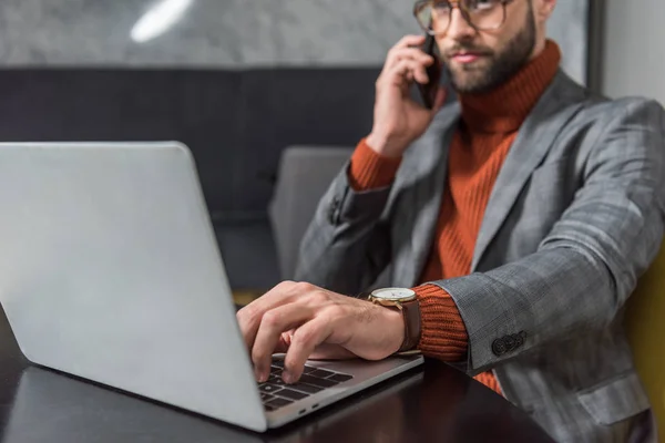 Vista cortada de empresário bonito em óculos e desgaste formal sentado à mesa, falando no smartphone e digitando no laptop no restaurante — Fotografia de Stock