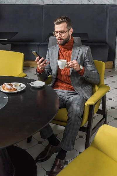 Hombre guapo en ropa formal usando teléfono inteligente mientras bebe café en el restaurante - foto de stock