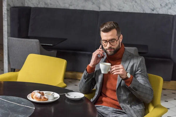 Focused handsome man in formal wear talking on smartphone while drinking coffee in restaurant — Stock Photo