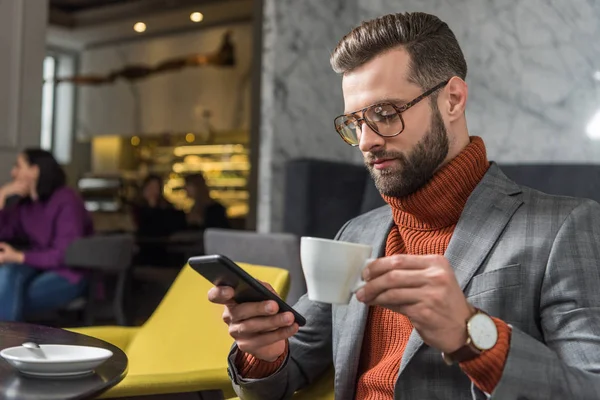 Concentré bel homme en tenue formelle et lunettes en utilisant smartphone tout en buvant du café dans le restaurant — Photo de stock