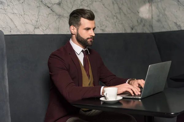 Bell'uomo d'affari in abito formale seduto a tavola con tazza di caffè e digitando sul computer portatile nel ristorante — Foto stock