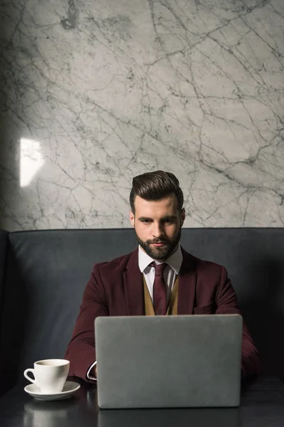 Серьезный привлекательный бизнесмен сидит за столом с чашкой кофе и печатает на ноутбуке в ресторане — стоковое фото