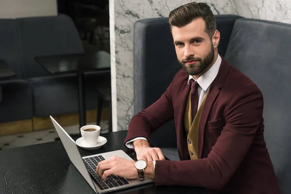 Hombre de negocios guapo mirando a la cámara, sentado en la mesa con una taza de café y escribiendo en el ordenador portátil en el restaurante - foto de stock