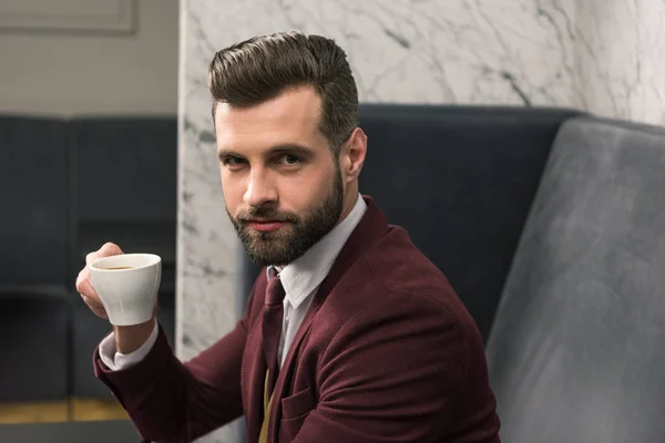 Handsome businessman in formal wear looking at camera, sitting at table and drinking coffee in restaurant — Stock Photo