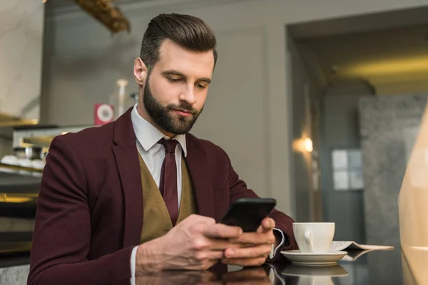 Hombre de negocios guapo en ropa formal sentado en la mesa con café y el uso de teléfono inteligente en el restaurante - foto de stock