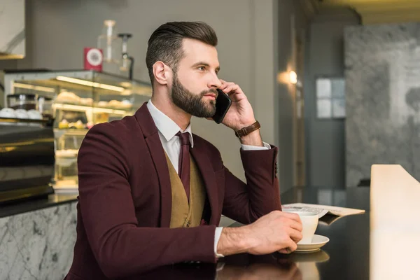 Серьезный привлекательный бизнесмен в формальной одежде сидит за столом и разговаривает по смартфону в ресторане — стоковое фото