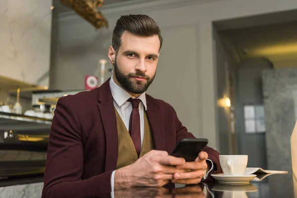 Sérieux bel homme d'affaires en tenue formelle assis à table avec du café et en utilisant smartphone dans le restaurant — Photo de stock