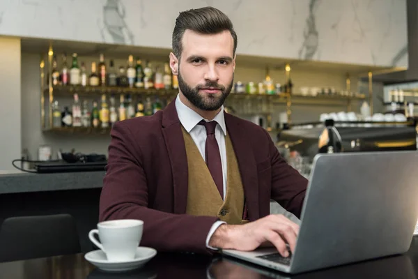 Homem de negócios bonito olhando para a câmera, sentado à mesa com xícara de café e digitando no laptop no restaurante — Fotografia de Stock