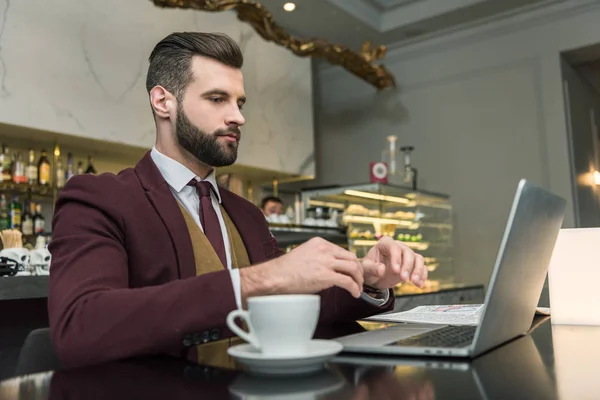 Serious handsome businessman sitting at table with cup of coffee and typing on laptop in restaurant — Stock Photo