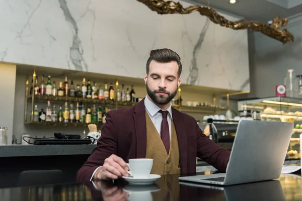 Bell'uomo d'affari che tiene una tazza di caffè e siede a tavola con un computer portatile nel ristorante — Foto stock