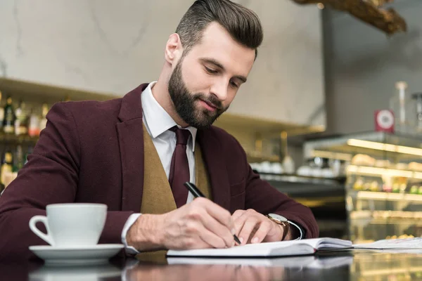 Guapo hombre de negocios en ropa formal sentado y escribiendo en cuaderno en la mesa en el restaurante - foto de stock