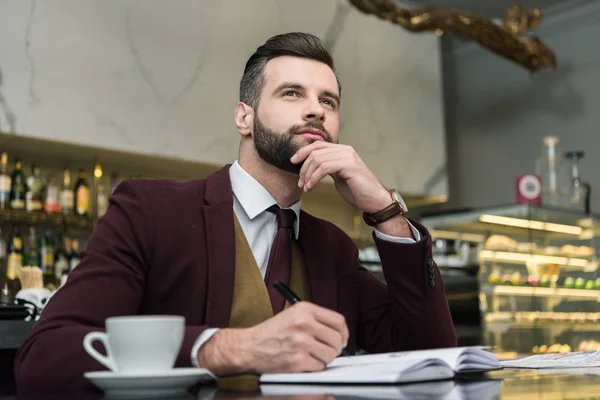 Nachdenklicher Geschäftsmann in formeller Kleidung sitzt und schreibt in Notizbuch am Tisch im Restaurant — Stockfoto