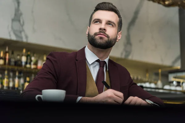 Thoughtful businessman in formal wear sitting at table and writing in restaurant — Stock Photo