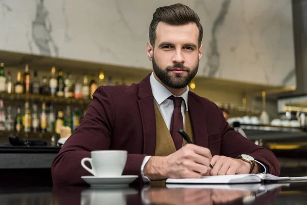 Homme d'affaires en tenue de cérémonie assis, regardant la caméra et écrivant dans un carnet à table dans un restaurant — Photo de stock