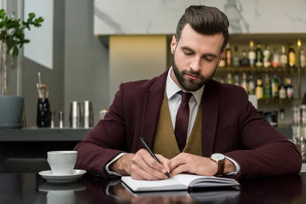 Serio hombre de negocios guapo en ropa formal sentado y escribiendo en el cuaderno en la mesa en el restaurante - foto de stock