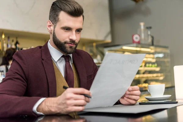 Handsome bearded businessman in formal wear sitting at table and looking at contract — Stock Photo