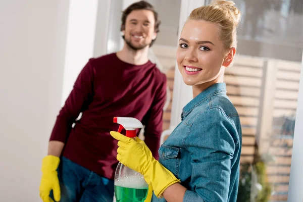 Menina bonita em luvas de borracha segurando spray com marido sorridente no fundo no apartamento — Fotografia de Stock