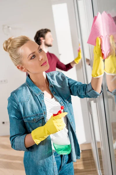 Beautiful girl cleaning window with rag and spray in apartment — Stock Photo