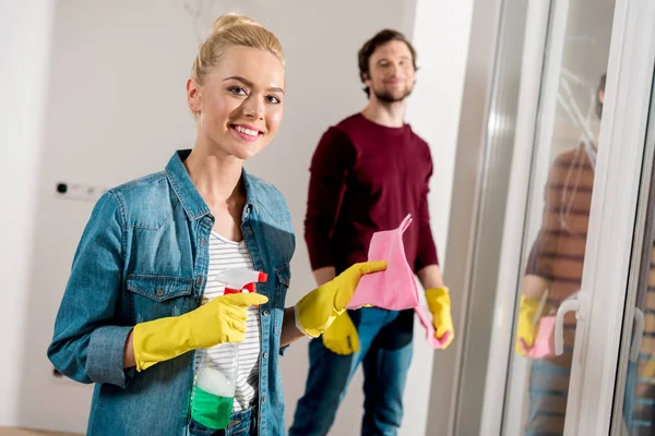 Attraktives Mädchen in Gummihandschuhen, das Spray und Lappen in der Hand hält, in die Kamera blickt und mit einem schönen Mann im Hintergrund lächelt — Stockfoto