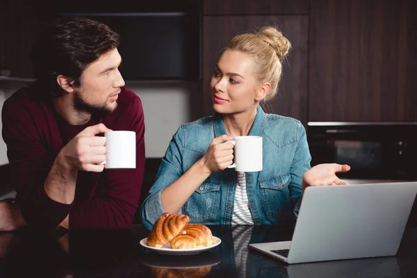 Menina atraente segurando xícara de café, apontando para a tela do laptop e olhando para o marido na cozinha — Fotografia de Stock